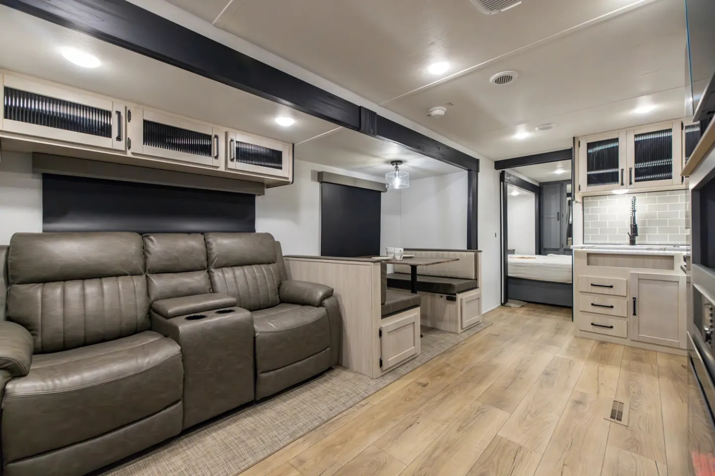 The living area of a travel trailer with a leather sofa, dinette, kitchen, and bedroom beyond